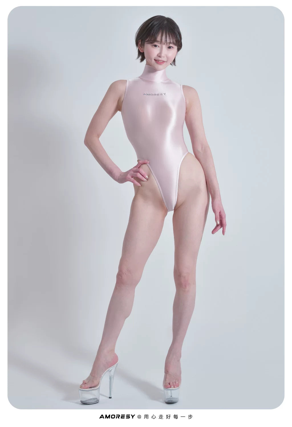A model wearing a shiny pale pink high-neck bodysuit with a small silver logo on the chest, paired with clear high-heeled platform shoes, standing against a light gray studio background.