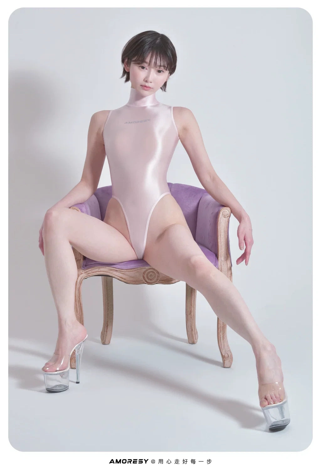 A model wearing a shiny pale pink bodysuit and clear high-heel platform shoes sits on a purple cushioned wooden chair against a light gray studio background.