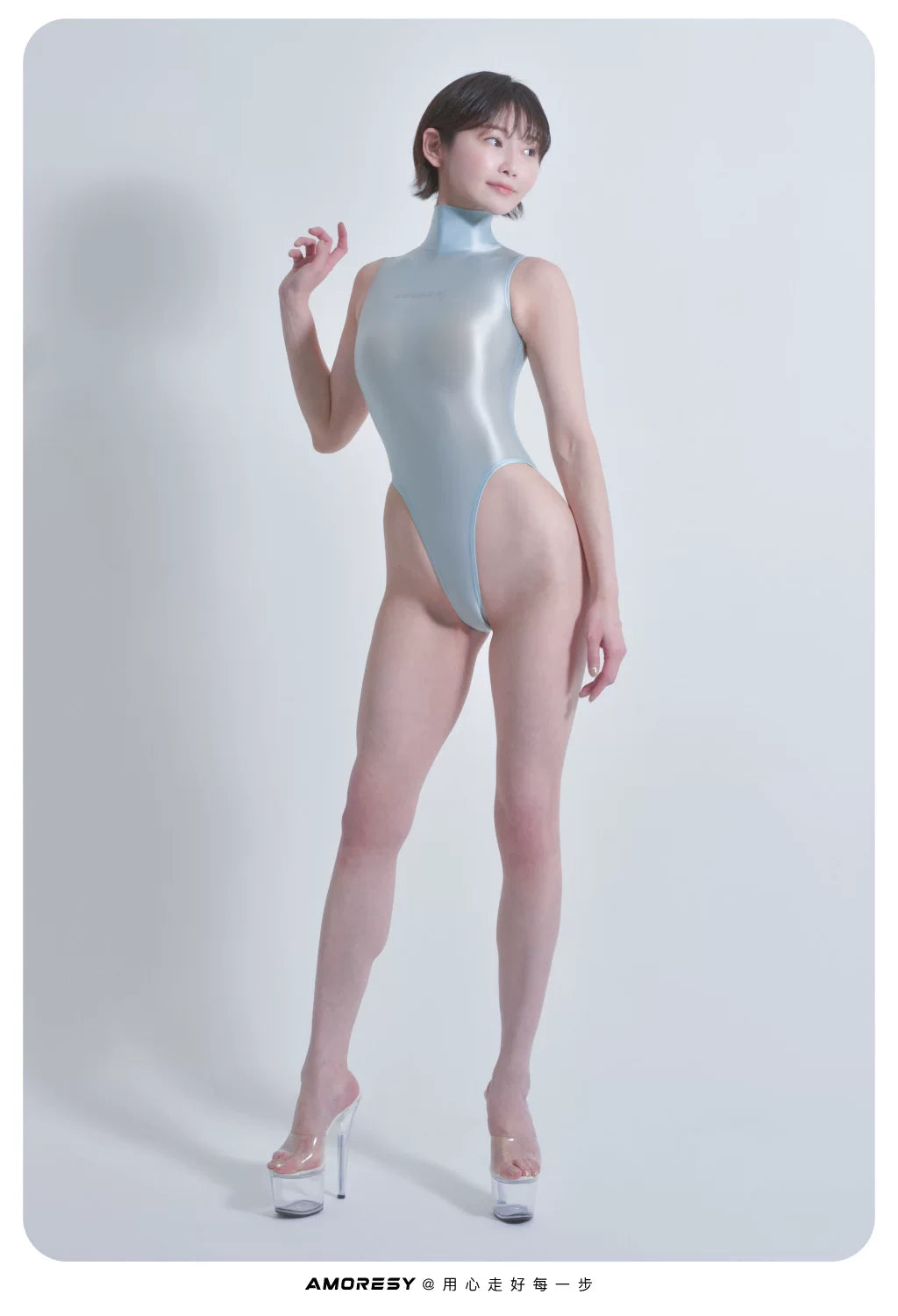 A model wearing a shiny, light blue high-neck latex bodysuit with a thigh slit, paired with clear high-heeled platform sandals, set against a plain light gray studio background.